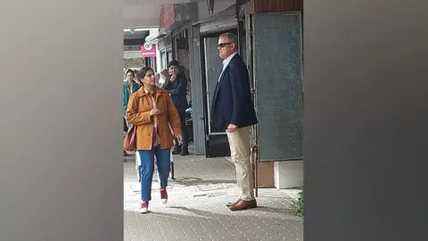 Kerry Evans Hugh Bonneville standing outside Gadebridge Chemist in Hemel during filming of Paddington in Peru. He is seen sideways on wearing sunglasses, a blue jacket and cream chinos, and brown shoes. He is pouting and clenching his fists as though slightly irate. People are in the background looking on, and one lady wearing a tan coloured coat and blue jeans is walking towards him - she might also be a cast member. 