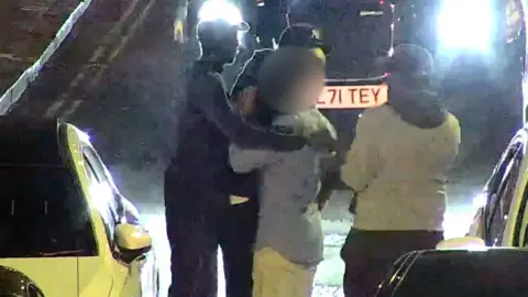 Two individuals distract victim in dark street, surrounded by parked cars and illuminated by headlights.