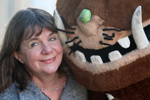 PA Julia Donaldson, a woman with shoulder-length dark hair, smiles while standing next to someone dressed in a Gruffalo suit.