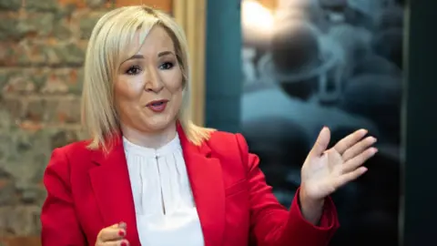 Michelle O'Neill, with shoulder length blonde hair, wearing a white top and a red suit jacket. She has one hand raised. 