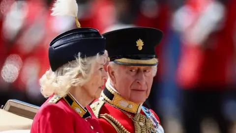 The King and Queen riding in a carriage. He is wearing the uniform of the Grenadier Guards as well as a number of medals. Queen Camilla is wearing a red, silk crepe, grenadier guards uniform dress and a black beret.