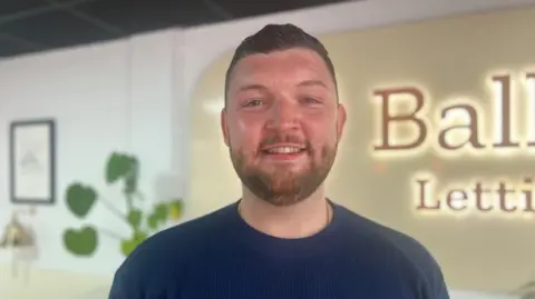 A man with short dark hair and a beard smiles at the camera. He is wearing a navy jumper, and is standing in front of a plant, picture and a sign for a lettings agency. 