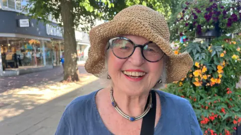 Phil Harrison/BBC A woman wearing a hat and a blue t-shirt is standing on a shopping street. She is looking at the camera and smiling.