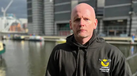 A man stands by a waterfront wearing a black hoodie branded with the UK Wildlife Crime Unit logo.