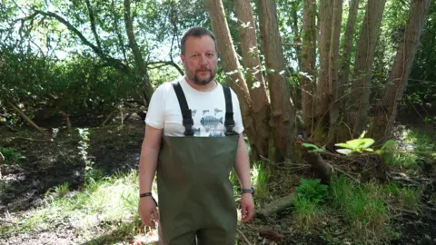 Shaun Whitmore/BBC Dr Jonah Tosney, who has a beard, is standing in the woodland wearing a white t-shirt and waders.