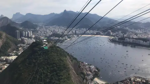 Courtesy of Leonardo Rocha A photo taken from Sugarloaf Mountain shows Morro da Urca and the bay below. The wires from the cable car can be seen linking the two mountaintops.
Highrise buildings rise in the distance.