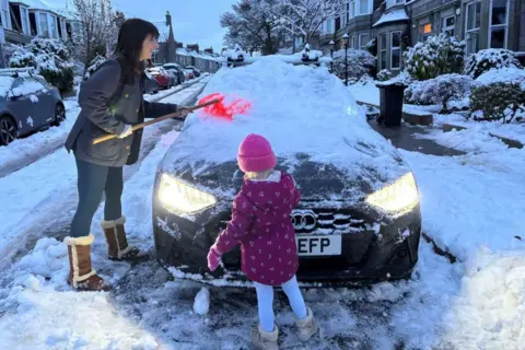 A mother and daughter clear the snow-laden windscreen of a car in the early hours. The car is on an upmarket residential street with sandstone properties, trees and bushes.
