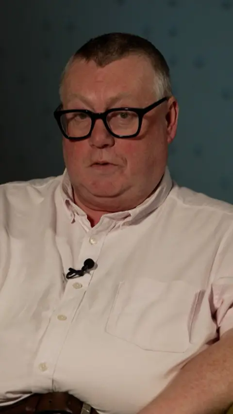 A man with short grey hair and black glasses sits in front of a greyish blue wall. He is wearing a light pink shirt and has a black lapel mic attached.