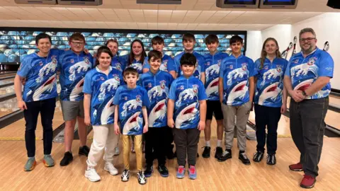 A group of 13 young bowlers alongside their coach. They are posing for a group photo in a bowling alley and smiling at the camera. They are all wearing matching blue shirts which depict a shark jumping out of waves. 
