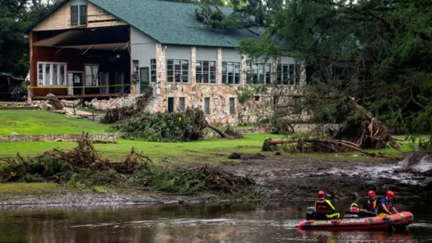People in a boat row down a river with a damaged building in the background and trees strewn over grass
