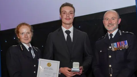 Wiltshire Police Ashton McCabe standing in the middle holding a small silver plaque. He is wearing a black suit and tie. A woman on his right is handing him a certificate. There is a man on his left who is wearing a row of medals on his chest.