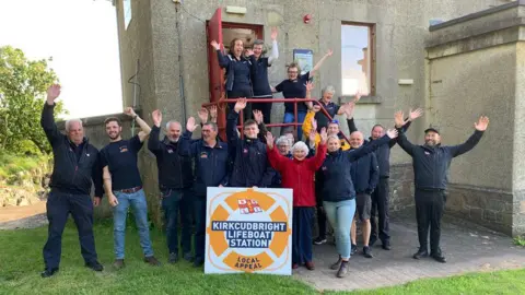 A group of people wave their hands in the air outside a lifeboat station to launch a local fundraising appeal