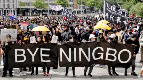 Reagan Yip A crowd of protestors holding a sign which reads 'Bye Ningbo'