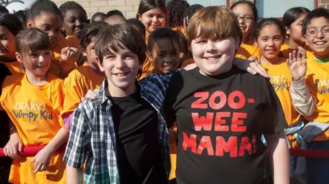 Getty Images Two children stand with their arms around each others' shoulders. Behind them are many more children wearing orange t-shirts with the "Diary of a Wimpy Kid" logo.