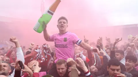PA Media Sam Hoskins in a pink Northampton Town shirt on the shoulders of a fan with an inflatable champagne bottle among a sea of supporters and smoke from a flare.