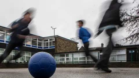 Three children in trousers are playing with a blue football. They are blurred and a school building is in the background.
