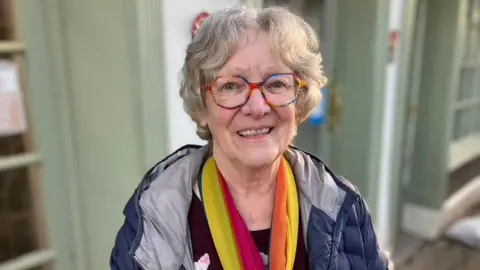 Wendy is standing in the street, wearing a colourful scarf and glasses.