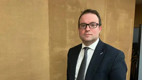 Amy Holmes/BBC Henry Vann looking into the camera with a textured brown wall behind him. He has short black hair and glasses, and is wearing a dark suit and tie over a white shirt.