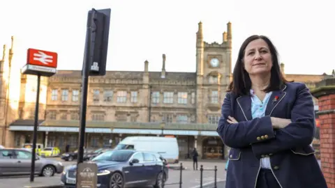 MP Julia Buckley, stood with Shrewsbury train station behind her. She is folding her arms, wearing a navy blue suit jacket and light blue shirt. There are cars in a car park behind.