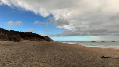 Brown sand dunes sit above the sand of a beach. The sea is to the right of the frame. A blue surfboard is at the bottom right of the frame, resting on the sand.