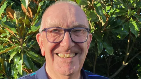 Man with glasses smiling at the camera with leaves of a plant behind him