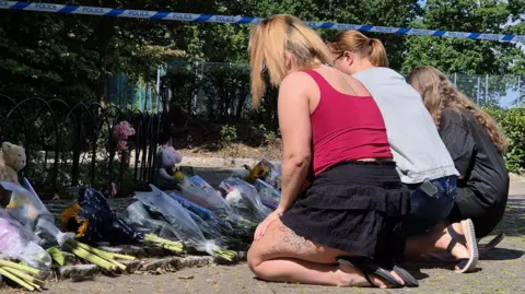 Stuart Woodward/BBC Three women kneeling in front of bouquets of flowers, which have been laid on the floor in a park. Behind the flowers is a fenced off bush.