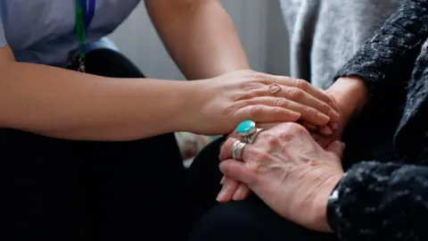 Two people holding hands. Only their forearms and hands are visible. The arm on the left belongs to a younger woman who has a diamond ring on her finger. The arm on the right of the image is covered by a dark knitted top and the person wearing it also has two rings on their fingers.