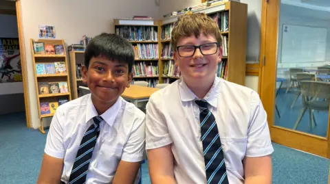 Ishrat is pictured on the left and Arlo is on the right. They are both wearing white short-sleeved shirts and a black, blue and white striped tie.