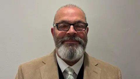 A man with glasses and shaved head and beard wearing a light brown jacket and shirt and tie