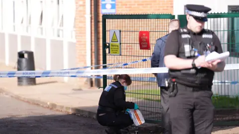A police officer standing by a green school gate holds up an evidence bag. She is wearing green plastic gloves and wearing a face mask. A police cordon separates her and a police officer standing in the foreground. 