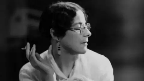 Black and white photo of Sylvia Townsend Warner smoking a cigarette. She is looking towards the right of the frame and holding the cigarette out to the left. She has dark hair, round glasses and pendant earrings.