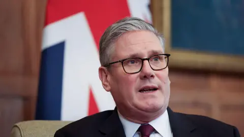 Sir Keir Starmer in a black suit and red tie. He is wearing glasses. The Union Jack is in the background.