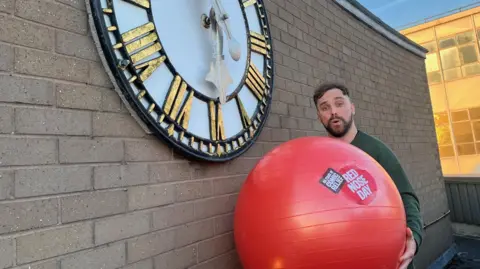 BBC Lee Blakeman, on the roof of the BBC Radio Stoke building, holding a large red medicine ball with Red Nose Day branding on it. He has short dark hair and a dark beard and stands next to a wall with a clock on it.