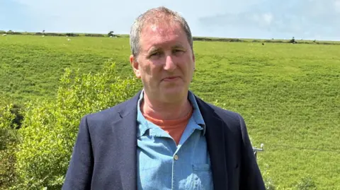 A man with grey hair wearing an orange t-shirt, blue shirt, and dark blue jacket standing in front of a green hill with sheep grazing on it.