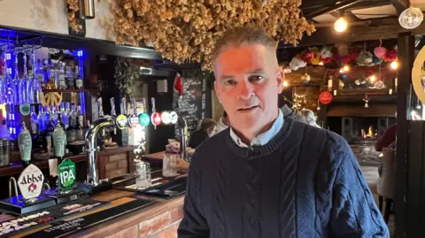 Phil Kiernan standing next to a row of pumps at a pub bar. He is wearing a navy knitted jumper and a light blue shirt with a collar