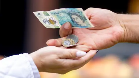 Getty Images A man's hand passes money to another person, including a £5 note and coins