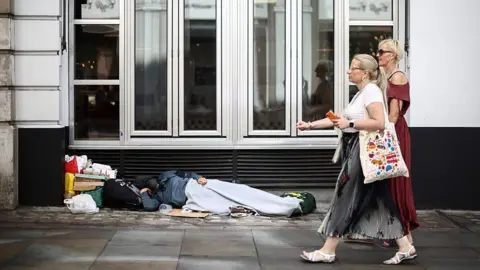 Getty Images A person lying covered by a grey blanket on cardboard outside a closed glass shopfront, surrounded by personal belongings and bags, while two pedestrians in summer clothing walk past on the pavement.