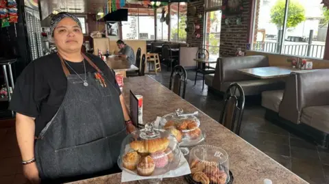 Raquel Pérez stands next to pastries in her restaurant. There is only one customer eating, and many empty tables