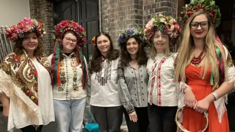 Six performers at the Ukrainian Christmas Service dressed in their national traditional costume during rehearsals. They have big floral head pieces, one woman is in a red dress and the other five have white or light coloured shirt-style tops with embroidered detailing. The woman in red is holding a tambourine.