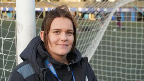 Tom Williams/BBC A woman wearing a dark jacket and identification lanyard standing in front of a soccer goal on a field, with blurred figures in the background.