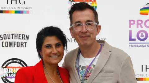 Getty Images Rebecca Paisis, wearing a red blazer and black top poses at a red carpet event with Simon Jones, who is wearing a floral t-shirt, blazer and glasses. They are stood in front of a board featuring Pride logos and adverts for sponsors. 