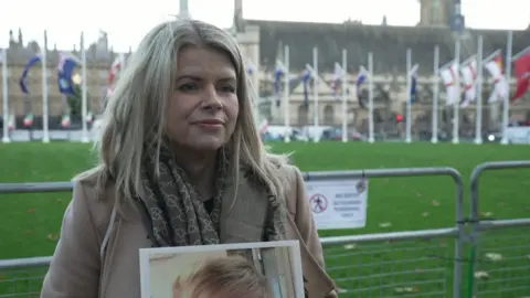 Louise Holmes standing in Parliament Square, London, holding a photo of her son and wearing a beige coat.