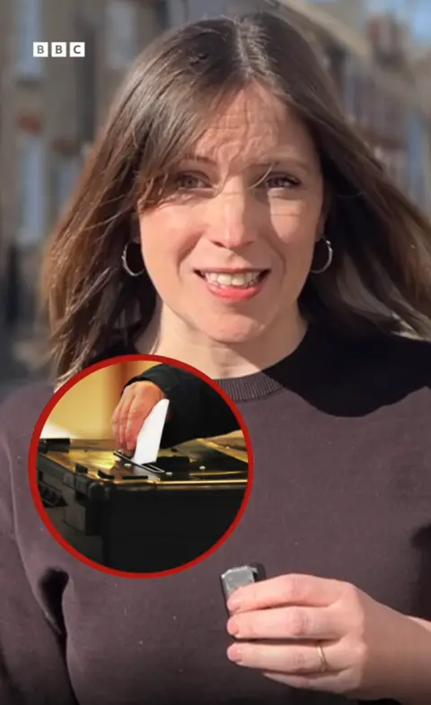 A reporter looks at the camera while talking. She is wearing a brown t-shirt and has short brown hair. There is a circular image of a person placing a ballot paper into a ballot box inset in the picture. 