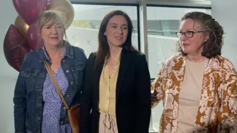 RTÉ Tori Towey in a suit jacket with yellow blouse, smiling beside a collection of pink and gold balloons that say welcome home Tori, at Dublin Airport - her mother is in an orange patterned top and beige shirt, her aunt in a blue patterned dress and denim jacket with brown leather crossbody bag