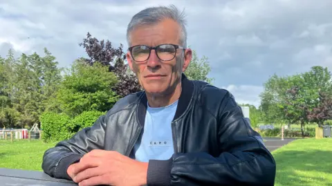 A man is sat at a park bench with his hands resting on the table, clasped in front of him. He has short dark hair which is turning largely grey. The man, dressed in a blue tshirt and black leather jacket, is looking at the camera through his large, round-rimmed glasses. Behind him is a footpath surrounded by grass on both sides, some trees and a children's play park in the distance. 