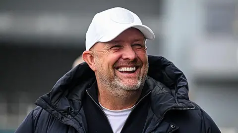 Andy Woodman, wearing a white cap and coat, laughs on the touchline