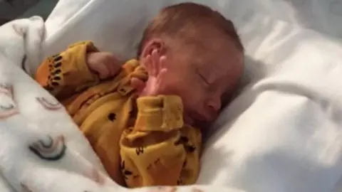 A newborn baby wearing a yellow sleep suit, laying in a bed covered with a blanket.