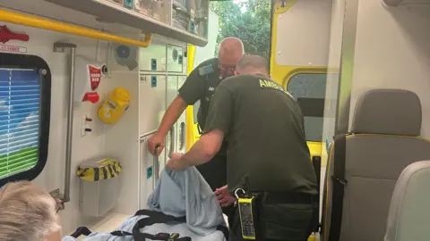 An elderly woman is lying on a cot in an ambulance being tended by two paramedics