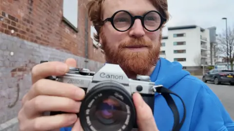 A man with ginger hair looks into the distant with his camera in hand wearing a blue jumper.
