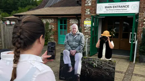 Prince of Wales School A child dressed as a classic jungle explorer stands next to a cut out of David Attenborough whilst a woman takes a picture with a phone.